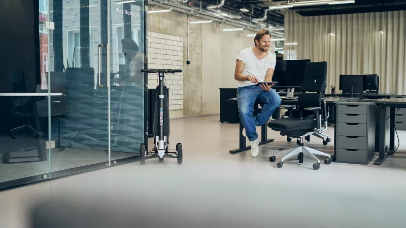 Man with a tablet sits in a modern office, with an XBoard e-scooter beside him.